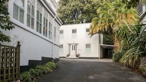 Een verharde oprit die leidt naar een gebouw met een witte gevel en meerdere ramen. Links bevindt zich een houten poort en rechts een garage.