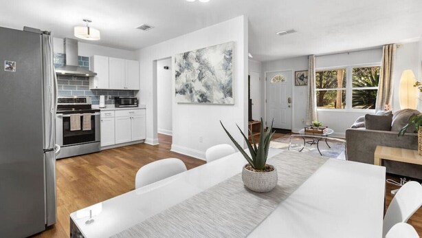 A modern kitchen with white cabinets, a stainless steel refrigerator, and a dining area with a white table and chairs. - Jeff GNV Home #1 · Renovated 3/2+ bonus, Heart of Gville, UF+Springs (Gainesville)