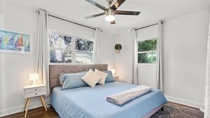 A bedroom with a bed, nightstands, a ceiling fan, and large windows. - Jeff GNV Home #1 · Renovated 3/2+ bonus, Heart of Gville, UF+Springs (Gainesville)