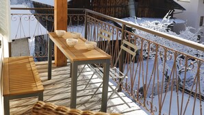 A wooden deck with a table and chairs, a wicker chair, and a metal railing overlooking a snowy landscape with buildings. - Tsaré Loft — Duplex 13 pers., Méribel (Les Allues)