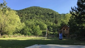 A wooden deck with a concrete edge, a small wooden playhouse, and a slide in a grassy area with a forested hill in the background. - Charming 3-star farmhouse  (Reilhanette)