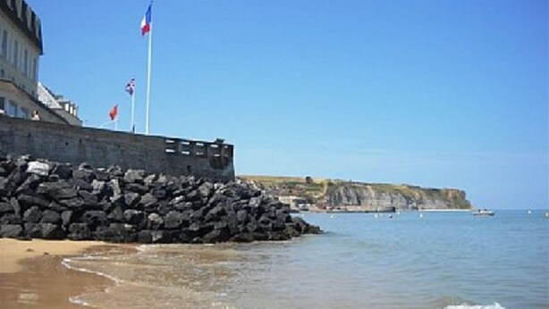 A coastal building with a stone wall and flags, overlooking a beach and cliffs.