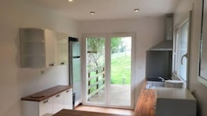 A modern kitchen with wooden flooring, white cabinets, and a large sliding glass door leading to a garden.