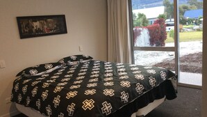 A bed with a black and white cross-patterned bedspread, a framed picture on the wall, and a view of a building and trees through a window. - Glenorchy rise (Glenorchy)