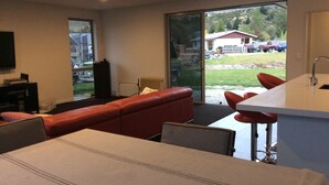 A modern living room with a sofa, a dining area with a bar, and a large sliding glass door leading to a view of a parking lot and buildings. - Glenorchy rise (Glenorchy)