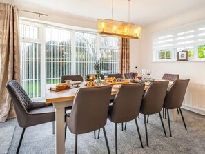 Uma sala de jantar com mesa posta para seis pessoas, rodeada por cadeiras, um candelabro e amplas janelas com cortinas.