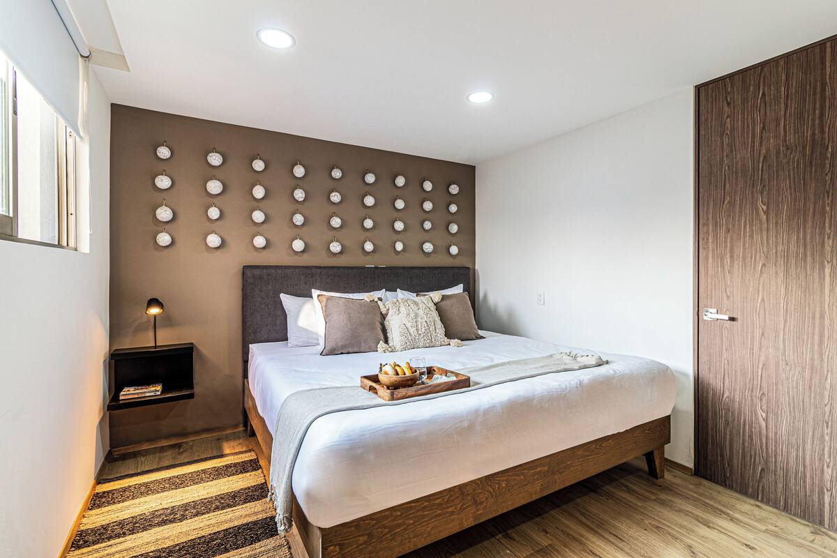 A modern bedroom with a large bed, a wall-mounted clock, a bedside table, and a wooden floor.