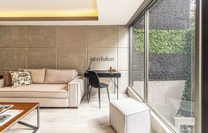 Een moderne woonkamer met een bank, een bureau en een groot raam met uitzicht op een tuin.