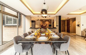 Een moderne eetruimte met een grote tafel gedekt voor een maaltijd, een keuken op de achtergrond en uitzicht door grote ramen.