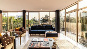 A modern living room with a dark blue sofa, a coffee table with books and a decorative item, and a view of greenery and buildings through large windows. - Casa Horacio by Kukun (Mexico City)