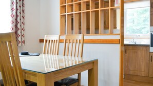 Un'area pranzo con un tavolo in vetro, sedie in legno, un divisorio in legno e vista su una cucina con mobili in legno.