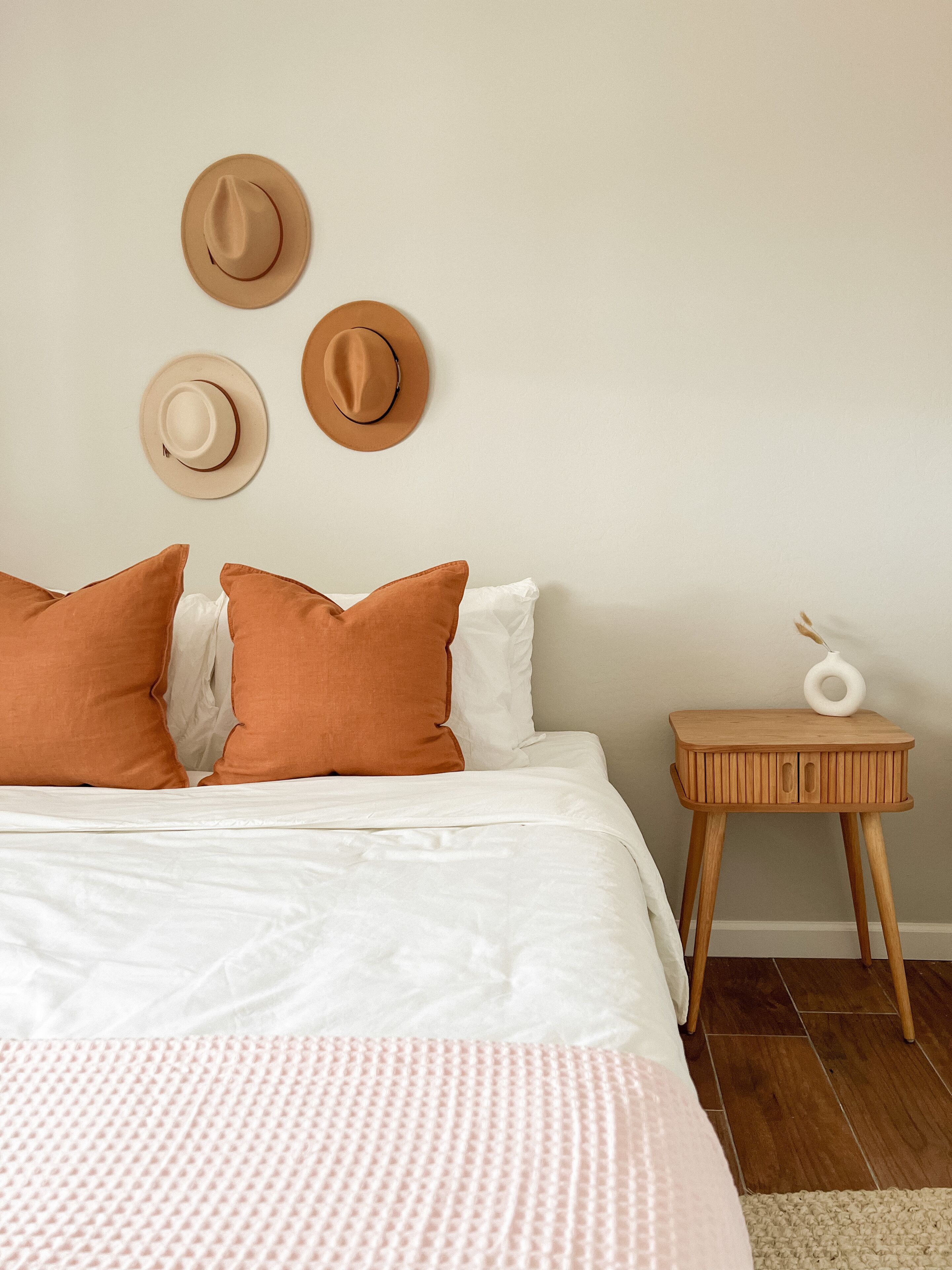 Ein ordentlich bezogenes Bett mit weißer und orangefarbener Bettwäsche, ein kleiner Nachttisch mit weißer Vase und drei Hüte an der Wand.