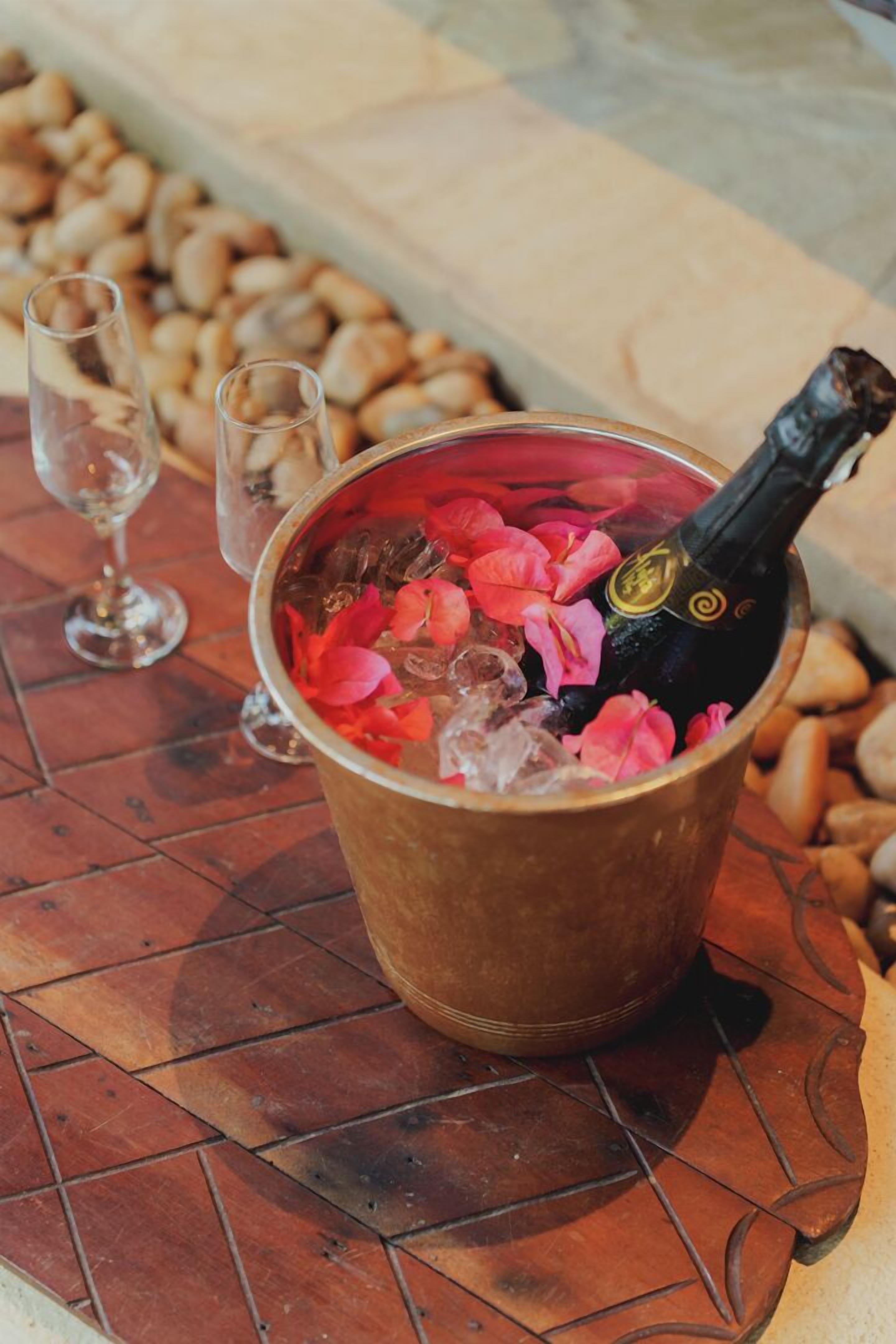 A bucket with ice, a bottle of champagne, and rose petals on a tiled surface.