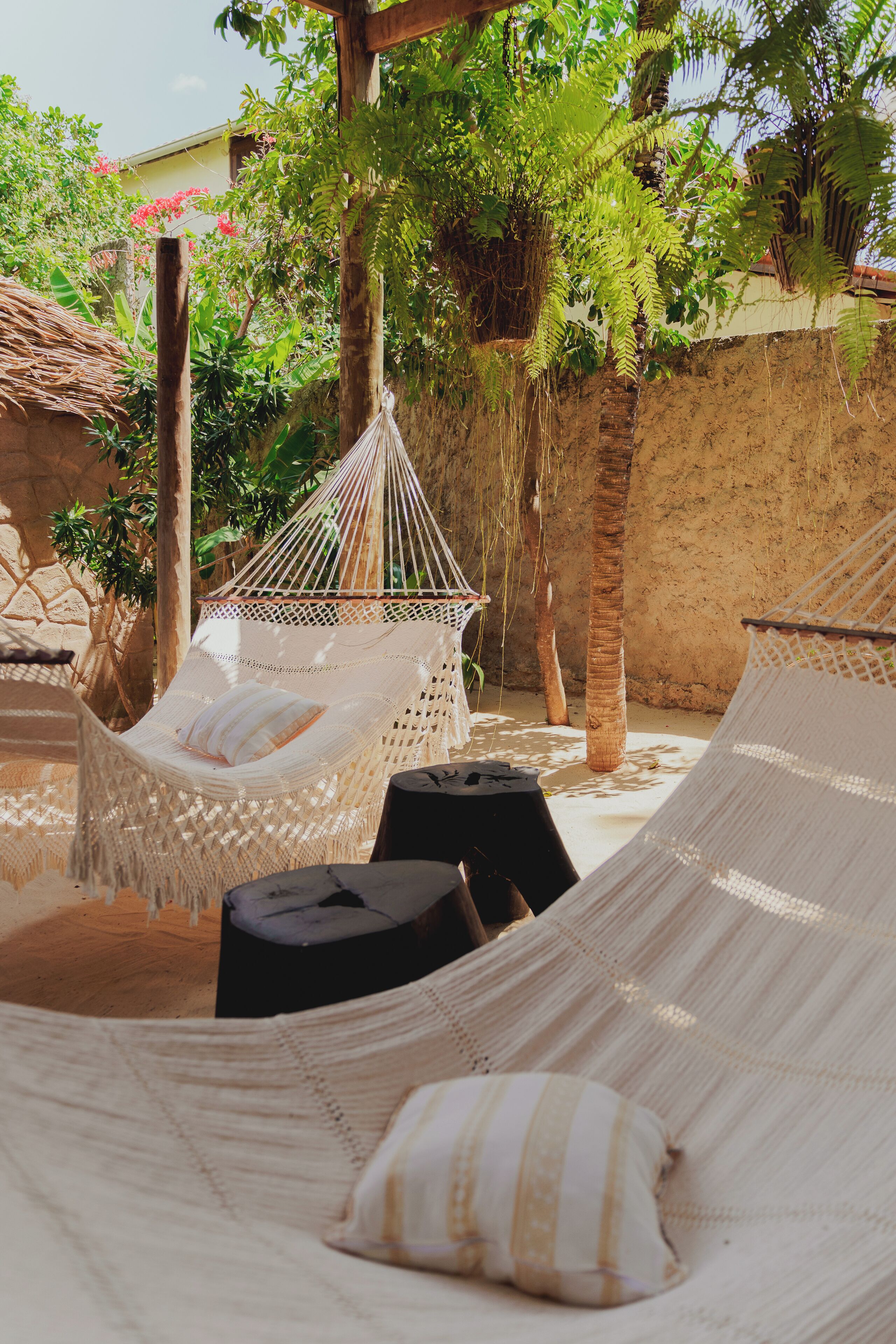 A hammock set up in a garden area with a stone wall and palm trees in the background.