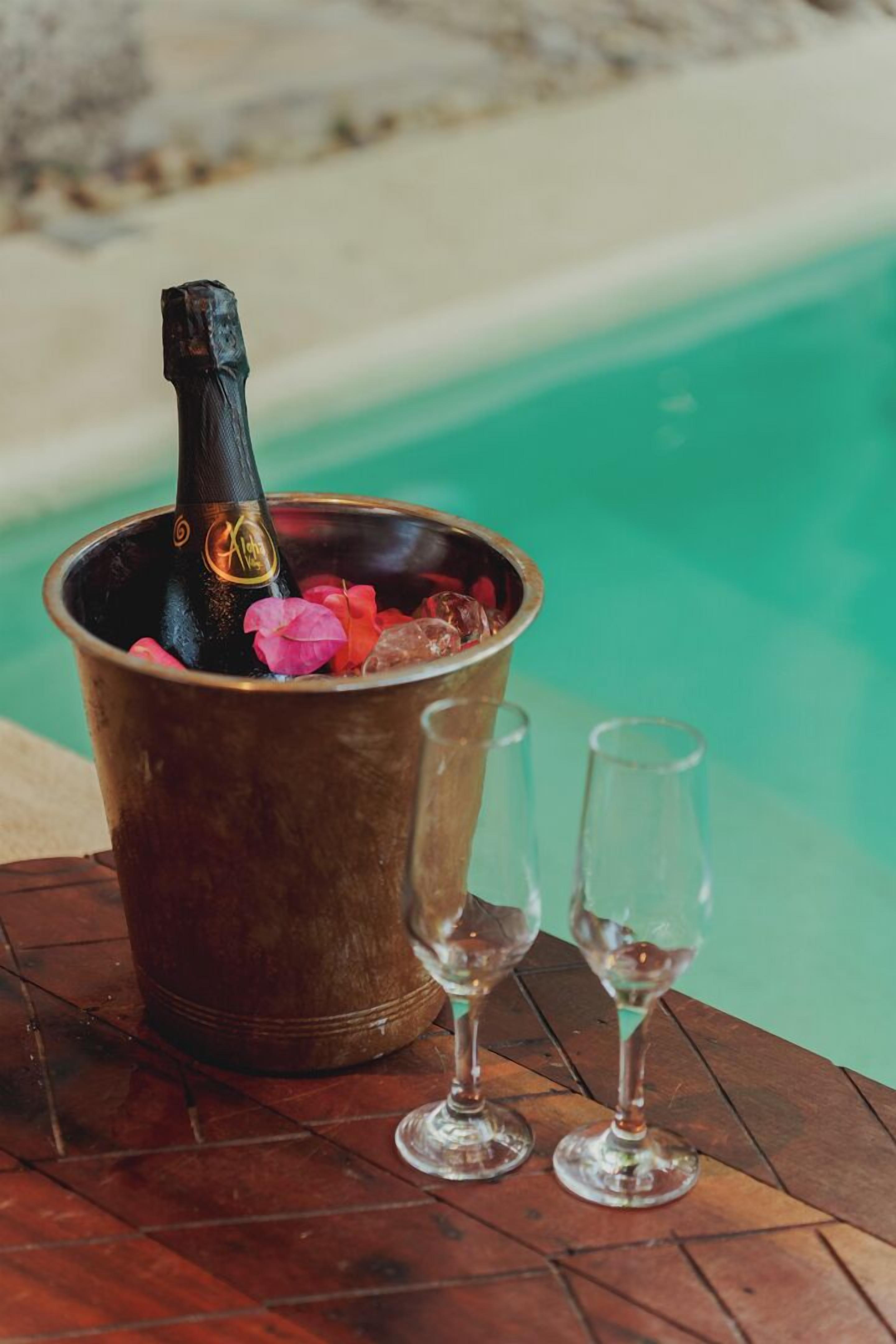 A bucket with a bottle of champagne and two glasses on a wooden table near a pool.