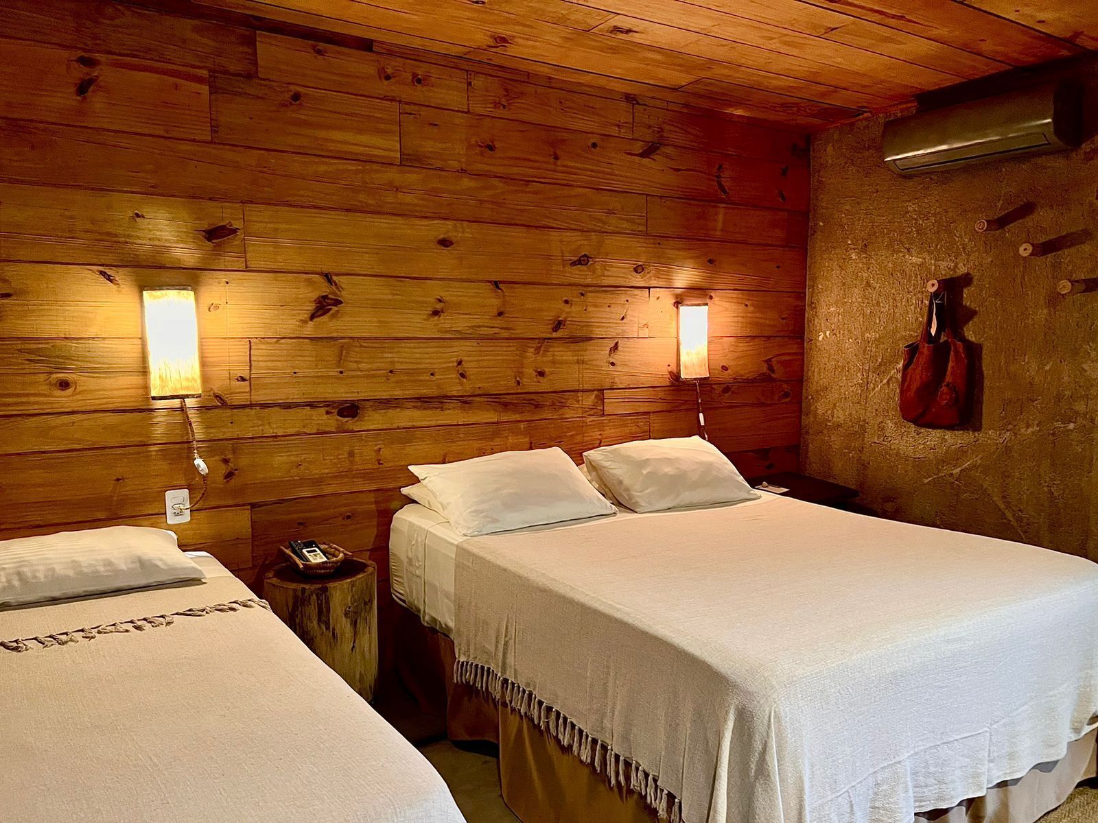 A wooden cabin room with two beds, each with white linens and pillows. There are wall-mounted lamps and a red bag hanging on the wall.