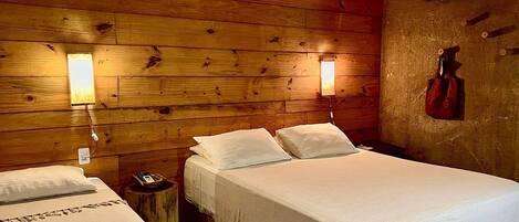 A wooden cabin room with two beds, each with white linens and pillows. There are wall-mounted lamps and a red bag hanging on the wall.