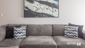 A gray sofa with two patterned pillows and a framed landscape painting of a snowy mountain. - New Charming Big Horn Ski-In/Out Home! Cozy Fireplace, Walk to Resort! (Big Sky)