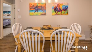 A dining area with a wooden table, white chairs, and two colorful paintings of bison on the wall. - New Charming Big Horn Ski-In/Out Home! Cozy Fireplace, Walk to Resort! (Big Sky)