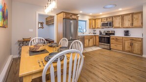 A modern kitchen with wooden cabinets, stainless steel appliances, and a dining area with a wooden table and white chairs. - New Charming Big Horn Ski-In/Out Home! Cozy Fireplace, Walk to Resort! (Big Sky)