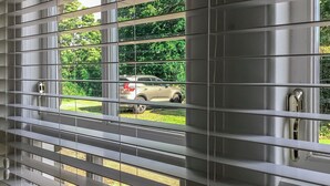 A window with horizontal blinds, a view of a parked car, and greenery outside. - 2 bedroom accommodation in Longhorsley, Near Morpeth (Longhorsley, Near Morpeth)