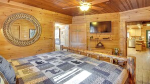 A cozy wooden cabin room with a large bed, a ceiling fan, a TV, and a view into the kitchen. - Hot Tub & Fire Pit: Riverfront Helen Cabin (Helen)