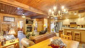 A cozy log cabin interior with a dining area, leather sofa, wooden table, and a kitchen with wooden cabinets. - Hot Tub & Fire Pit: Riverfront Helen Cabin (Helen)