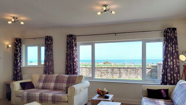 A living room with a sofa, armchairs, a coffee table, and a view of the sea through large windows. - 3 bedroom accommodation in Beadnell (Beadnell)