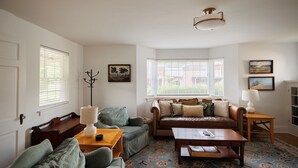 A cozy living room with a sofa, armchairs, a coffee table, and a fireplace. There are two windows with blinds, a ceiling light, and a rug on the floor.