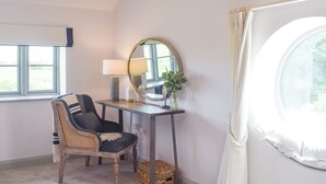 A room with a wooden chair, a small table with a lamp, a mirror, and a window with curtains.
