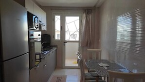 Een kleine keuken met een koelkast, oven en magnetron. Er is een eettafel met stoelen en een raam met zonwering.