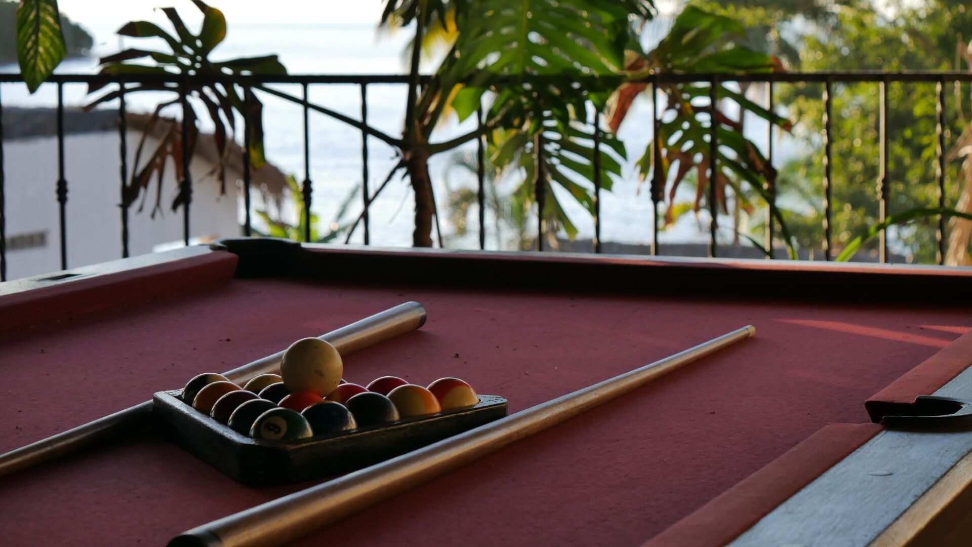 A pool table with balls and cues, set against a balcony with a view of the sea and greenery.