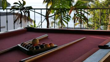 A pool table with balls and cues, set against a balcony with a view of the sea and greenery.
