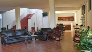 A spacious living area with a leather sofa, a coffee table, and a rocking chair. A staircase with red steps leads to an upper floor. The kitchen has wooden cabinets and a dining table. - Casa Rinconsito Miramar (Santa Cruz)