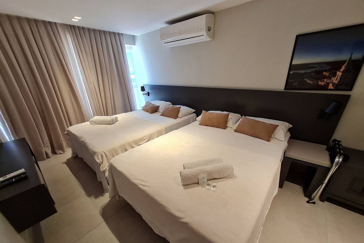 A hotel room with two beds, a flat-screen TV, an air conditioner, a painting, and a view of a cityscape.