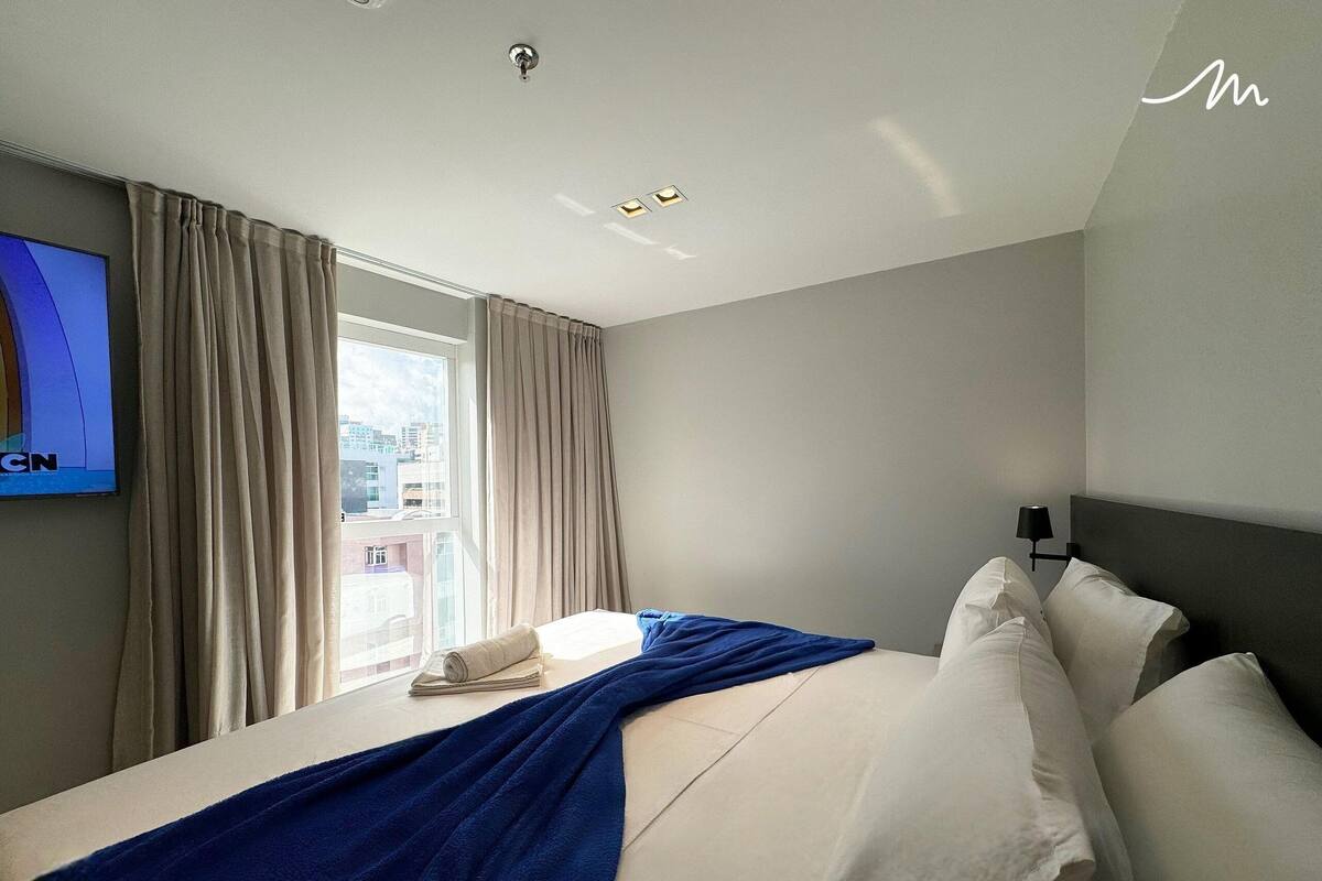 A hotel room with a bed, a TV, a window with curtains, and a view of buildings outside.