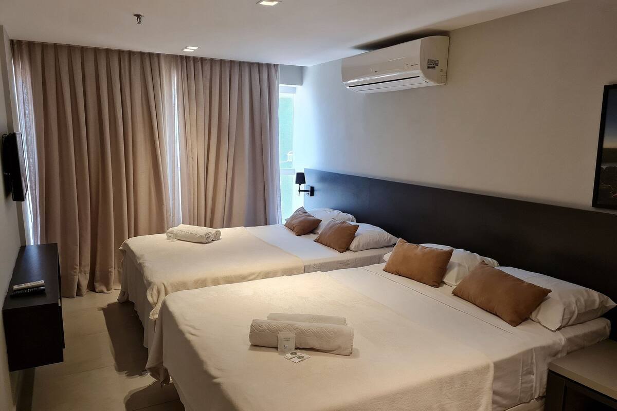 A hotel room with two beds, beige curtains, an air conditioning unit, and a flat-screen TV.