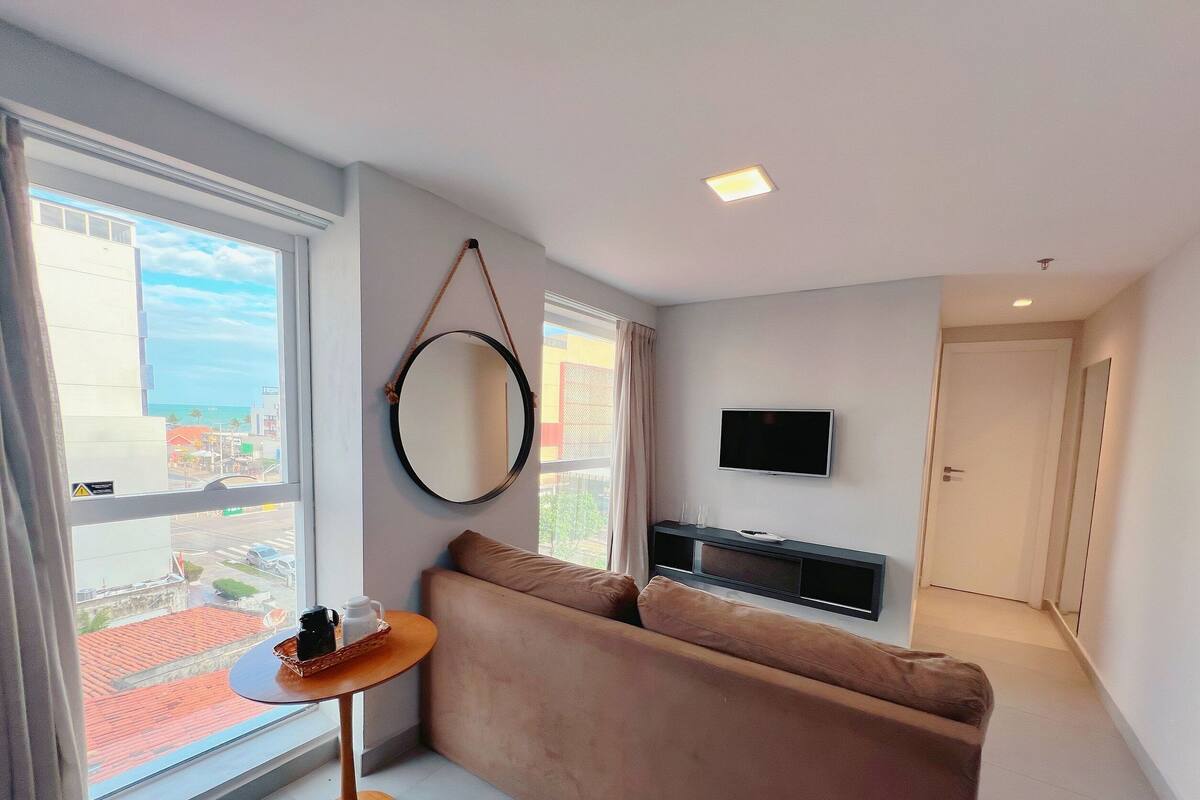 A modern living room with a sofa, a small table with a coffee pot, a TV mounted on the wall, and a large window offering a view of the city.