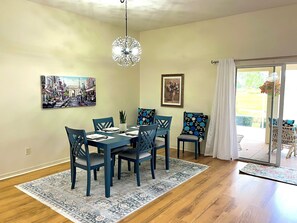 Uma sala de jantar com uma mesa posta para quatro pessoas, rodeada por cadeiras azuis, um tapete estampado e duas pinturas penduradas na parede.