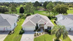 A residential area with houses featuring a central building with a flag, surrounded by well-maintained lawns and trees. - Golf View in the Villages (The Villages)