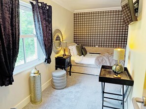 A bedroom with a bed, a bedside table with a lamp, a mirror, a window with curtains, and a checkered wall.