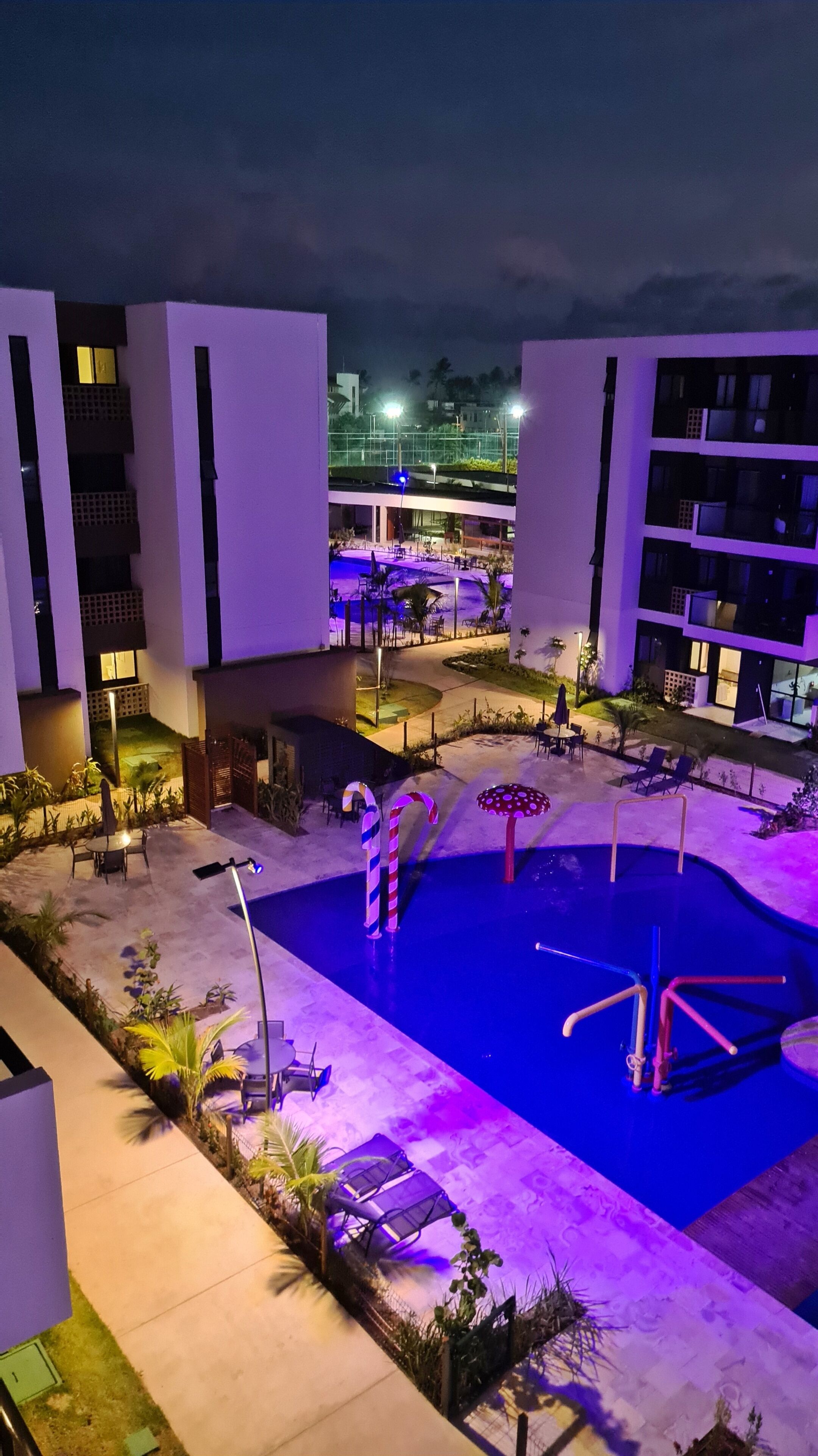 A pool area with lounge chairs and umbrellas, surrounded by buildings with balconies and a view of a distant cityscape.