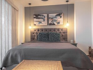 A bedroom with a tufted headboard, two abstract paintings, a bed with a grey quilt, a woven rug, and a small statue on the nightstand.