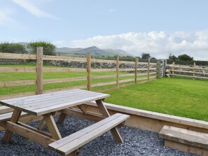 A picnic table with benches, surrounded by a stone wall and a wooden fence, overlooking a grassy field with mountains in the distance.