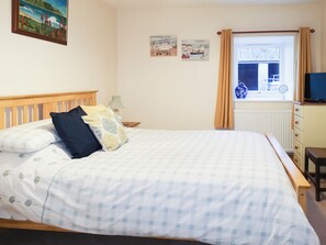 A bedroom with a bed, a nightstand, a lamp, a dresser, a television, and a window with curtains.