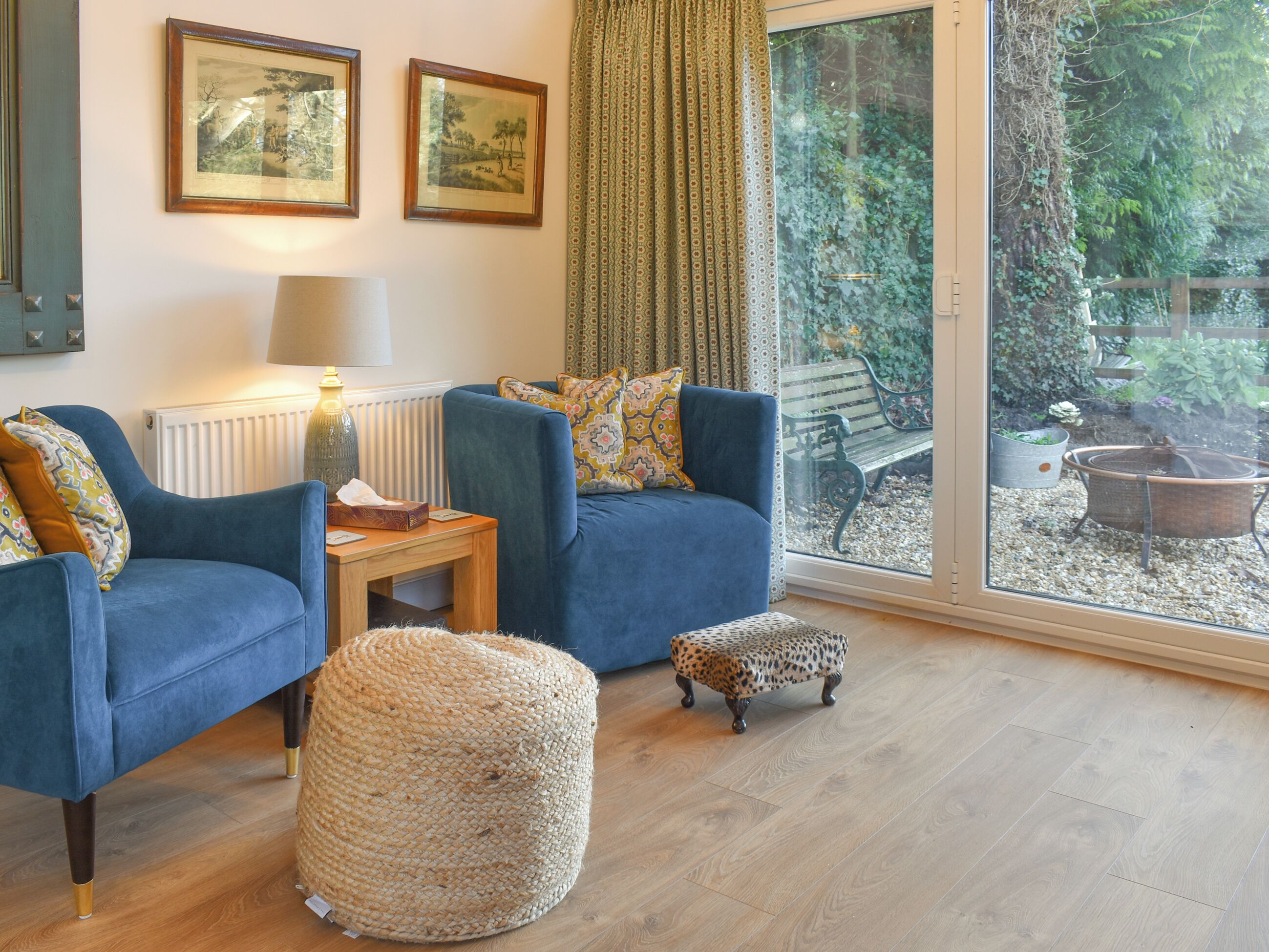 A living room with two blue armchairs, a small round ottoman, a wooden side table, and a floor lamp. There are two framed pictures on the wall, a window with a view of a garden, and a radiator under the lamp.