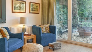 A living room with two blue armchairs, a small round ottoman, a wooden side table, and a floor lamp. There are two framed pictures on the wall, a window with a view of a garden, and a radiator under the lamp.
