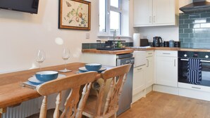 A modern kitchen with white cabinets, a wooden dining table with chairs, a flat-screen TV, and a window with blinds.
