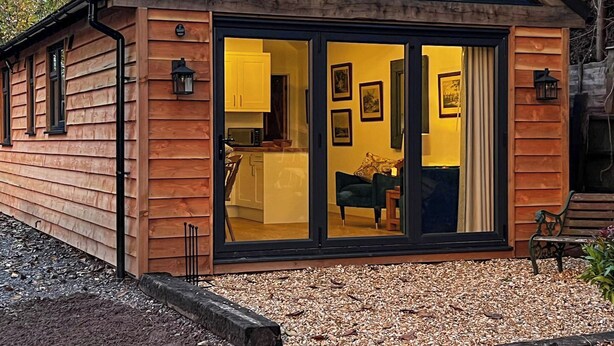 A wooden cabin with a large glass door, a small kitchen visible inside, and a bench outside.