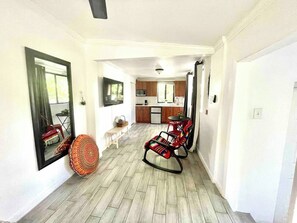 A spacious living area with a wooden floor, a rocking chair, a small table, and a kitchen in the background.